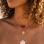 Woman wearing Seravelle Necklace, a layered gold necklace with red bead accents, paperclip chain, coin charm, and shell pendant