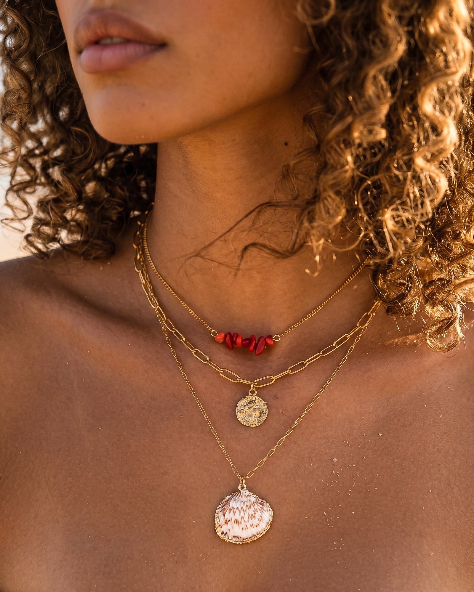 Woman wearing Seravelle Necklace, a layered gold necklace with red bead accents, paperclip chain, coin charm, and shell pendant