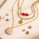 Seravelle Necklace displayed on a stone background with layered gold chains, red beads, and shell pendant