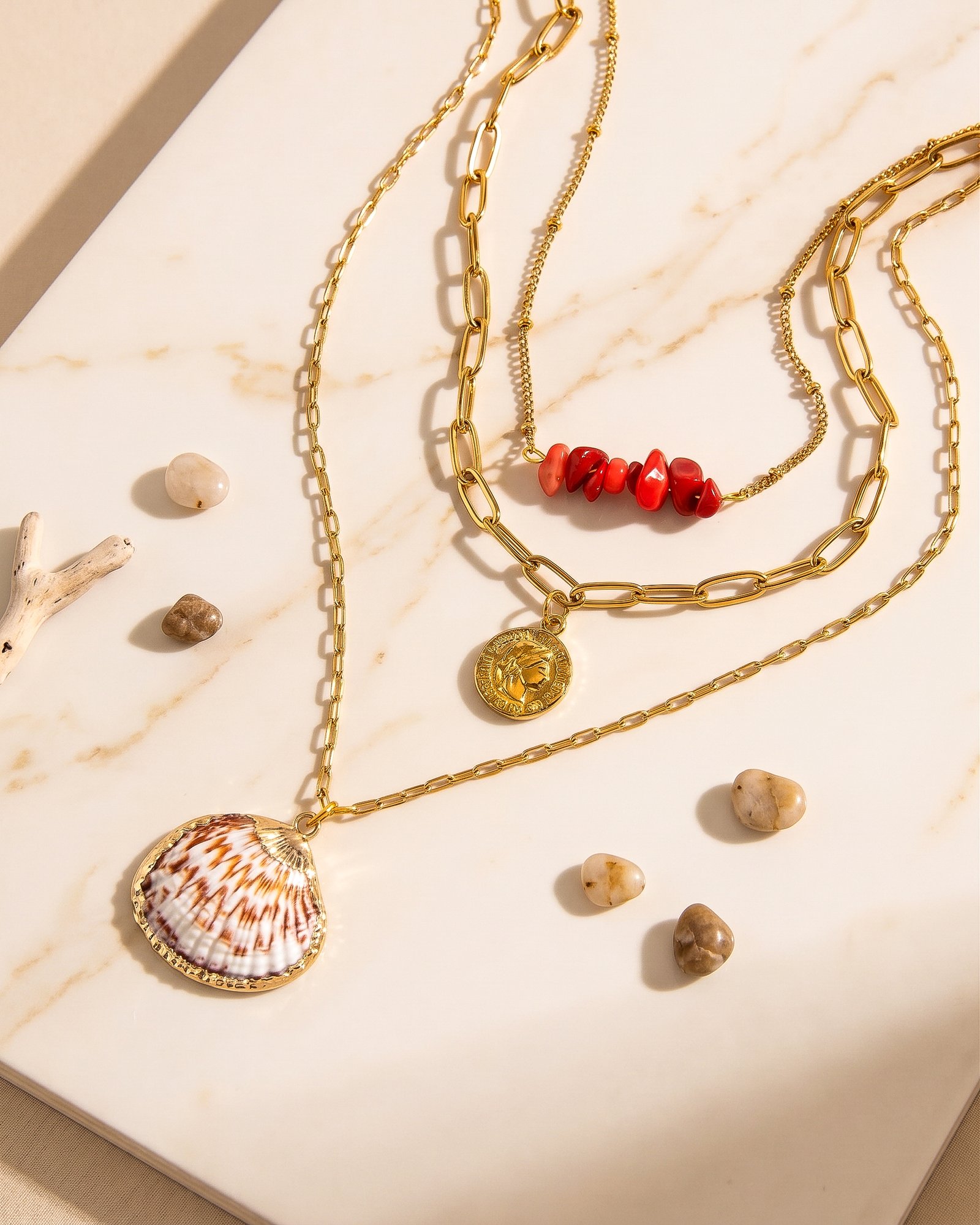 Seravelle Necklace displayed on a stone background with layered gold chains, red beads, and shell pendant